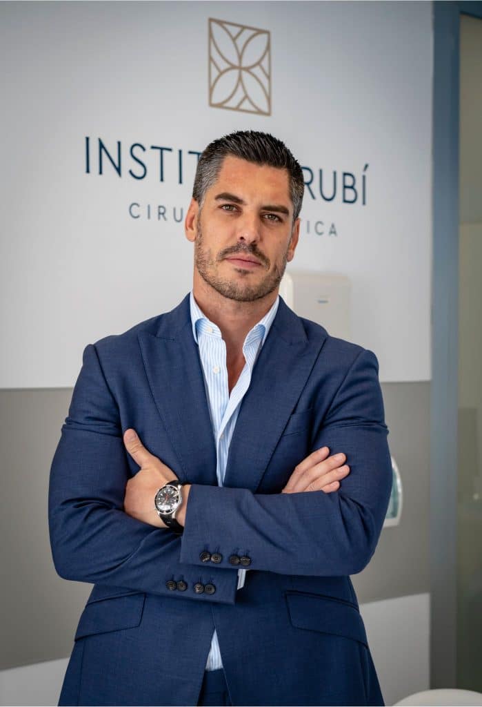 Diseño sin título (64) cirujano plástico en Instituto Rubí con traje azul, posando con los brazos cruzados frente al logotipo de la clínica
